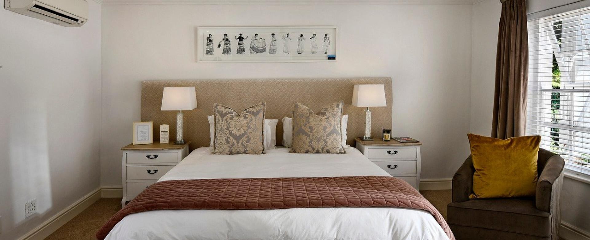 Unterkünfte Hotelzimmer, mittig ein mit weißer Bettwäsche und braunem Teppichüberwurf bezogenes Bett, beiges Kopfteil, hellen Lampen rechts und links auf weißen Nachttischen, an der Decke ein Deckenventilator, links eine Klimananlage an der Wand, rechts ein Fenster, davor ein Sessel