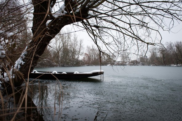 Winterwanderung Stocherkahn, mit Schnee bedeckt, liegt am Ufer des Gießen, im Vordergrund ein kahler Baum, dessen Stamm teilweise mit Schnee besprenkelt ist, im Hintergrund der graue Winterhimmel