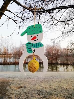 Futterstation Schneemann Vor dem Hintergrund des Sees hängt an einem winterlichen Ast ein Schneemann aus Holz, der bemalt ist und unten eine Aussparung vorhanden ist, in die ein Meisenknödel eingehängt werden kann