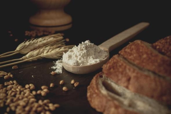Brotbacken auf einer schwarzen Unterlage leigen Getreideähren, -körner und ein Holzlöffel mit Mehl, im rechten Vordergrund mehrere Brotscheiben